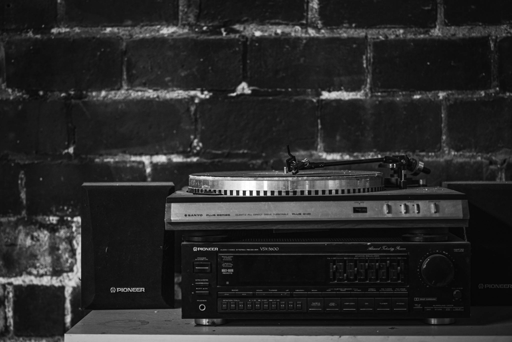 Monochrome image of a vintage turntable and audio setup with speakers against a brick wall.